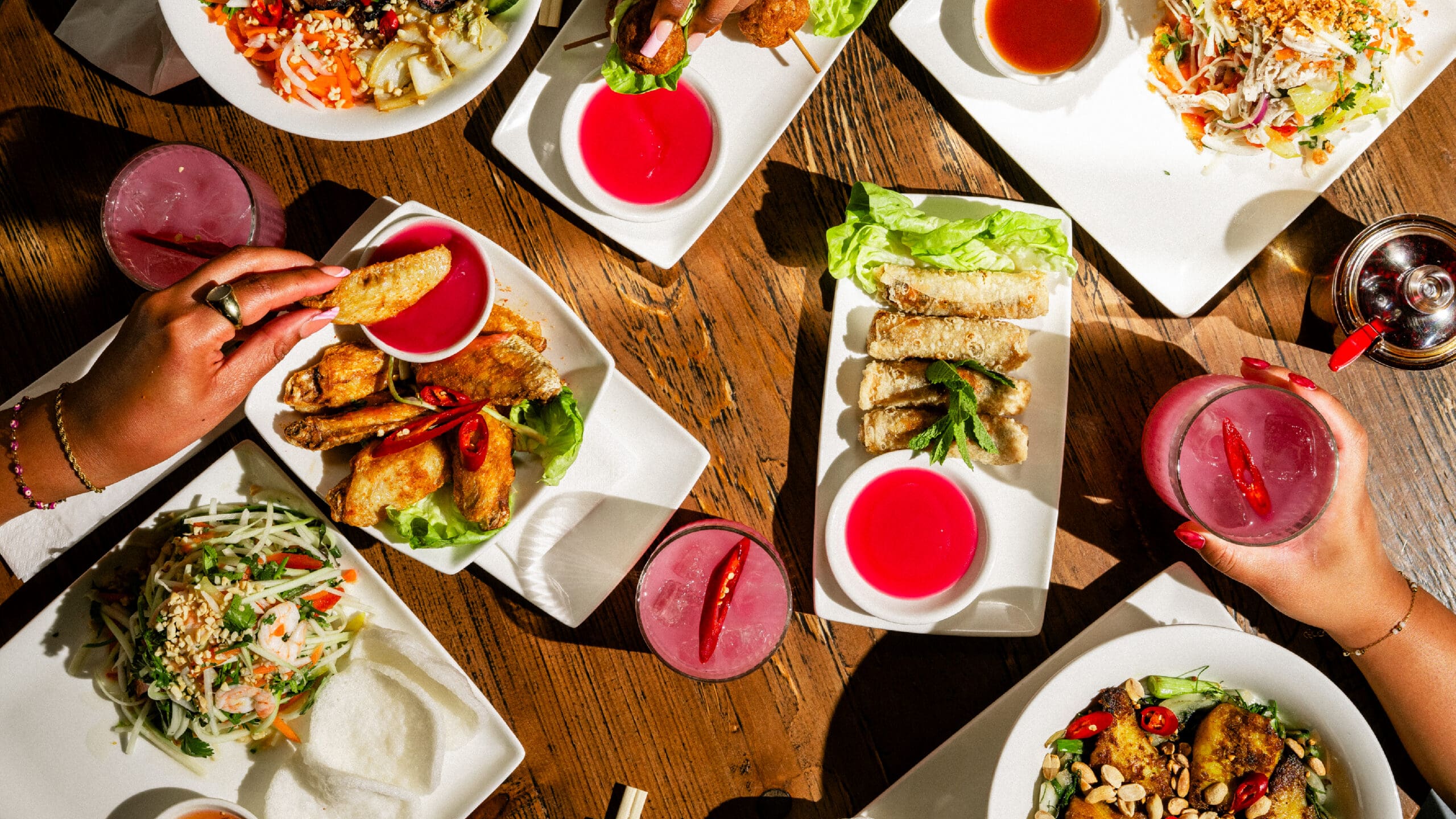 A table set with Vietnamese dishes, including spring rolls, salads, fried appetizers, and pink drinks. Two hands are reaching for food and drinks. The setting is bright, with sunlight illuminating the wooden table.