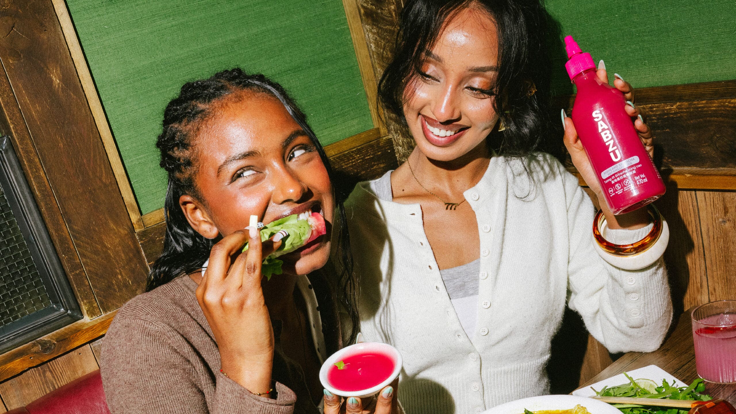 Two women sit at a table. One is about to bite a taco, dipping it in pink sauce. The other smiles, holding a bright pink squeeze bottle labeled “Spabzu.” Plates of food and drinks are on the table.