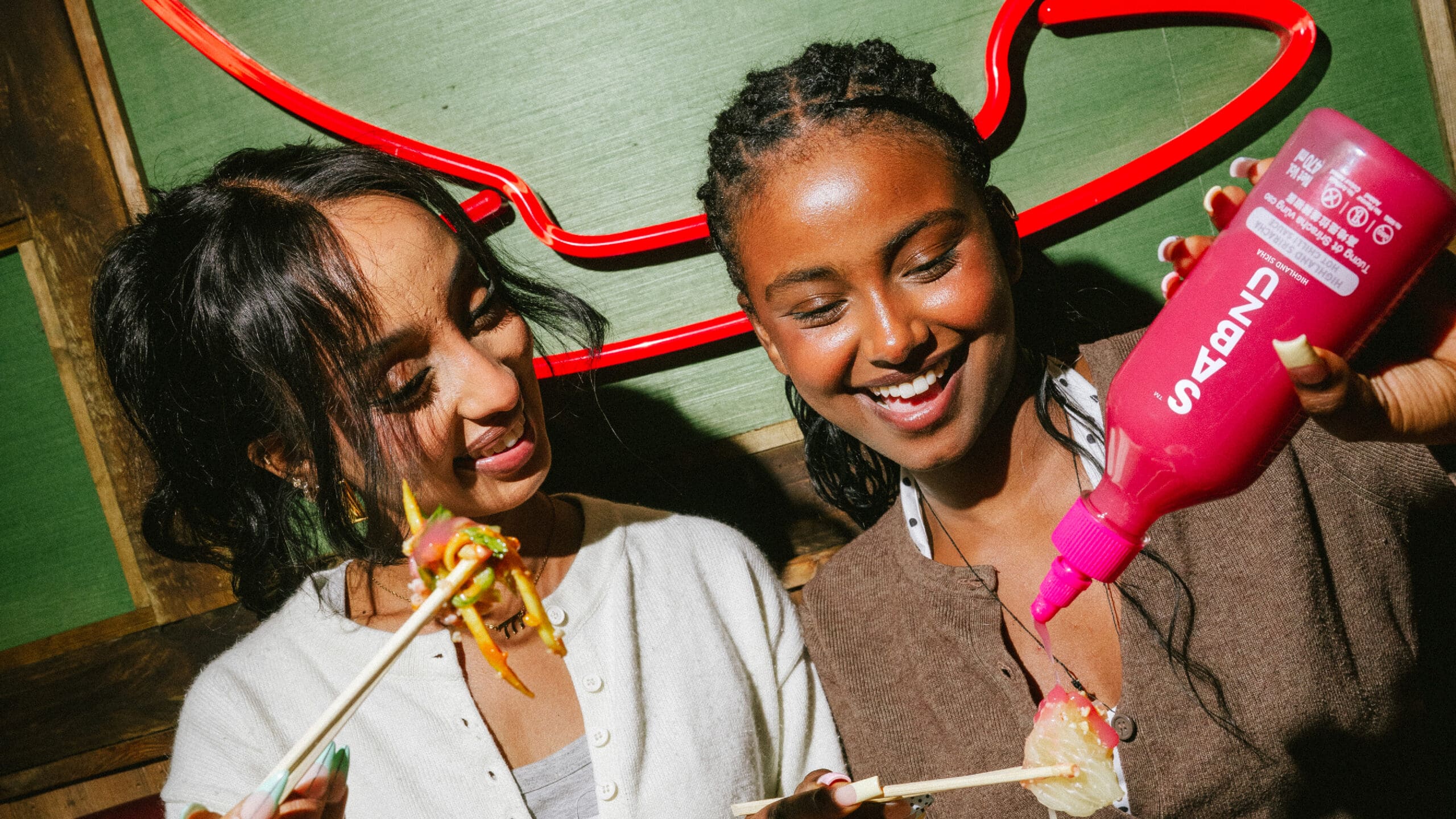 Two women smiling while eating with chopsticks; one pours sauce from a large pink bottle onto food. Neon red decor glows behind them, creating a lively, casual atmosphere.