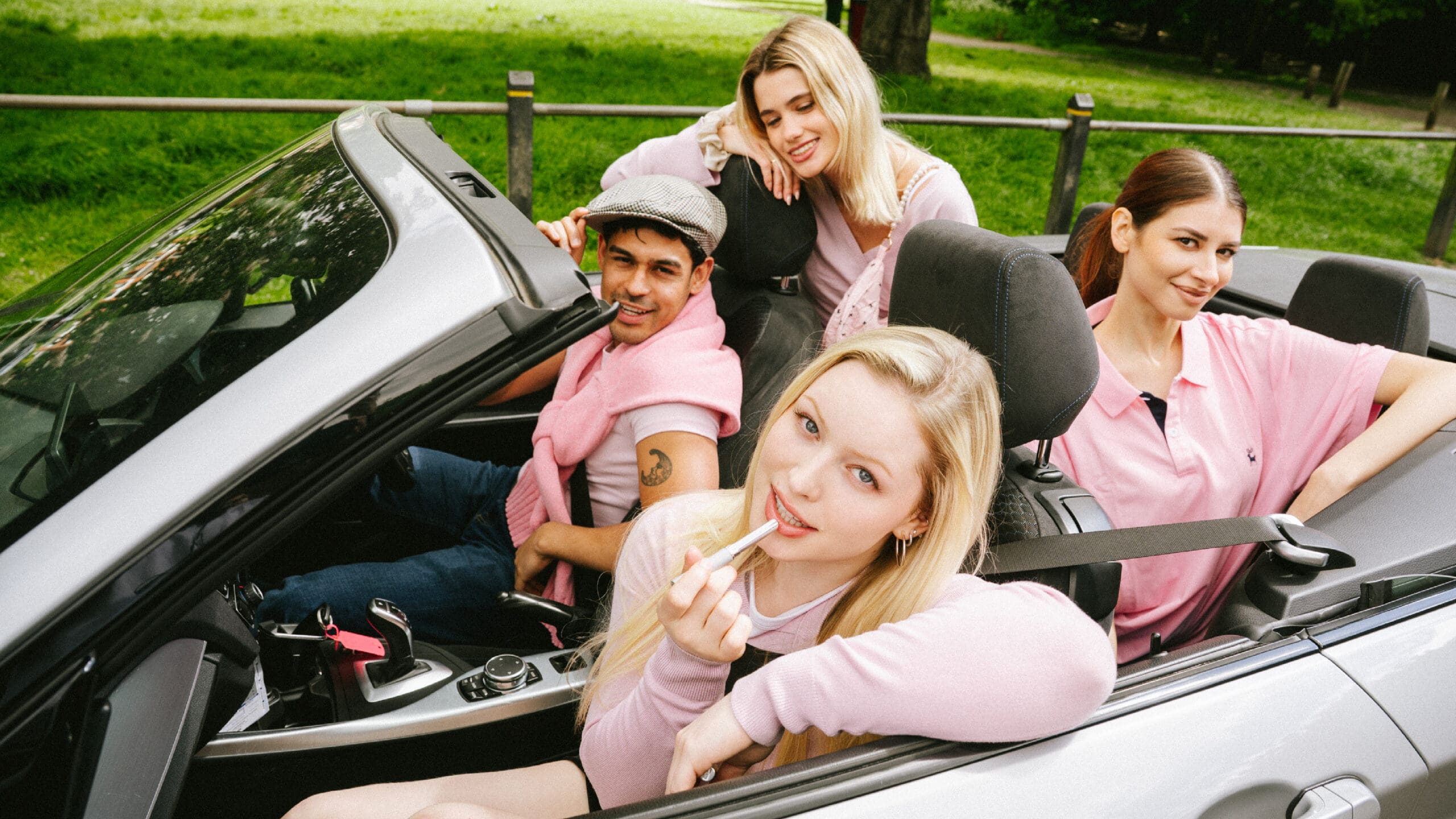 Four young adults wearing pink clothing sit in a convertible car with the top down, smiling and posing for the camera in a green park setting. One person is applying lipstick.