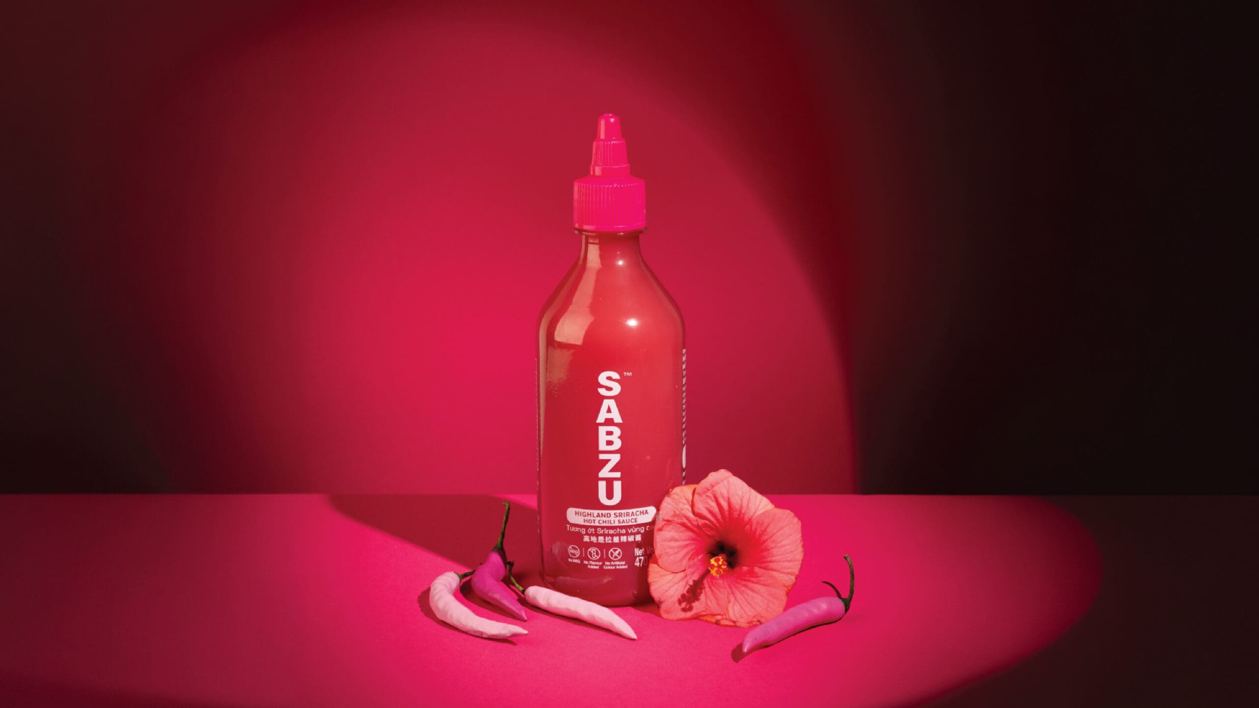 A red bottle labeled “SABZU” stands on a pink surface with three chili peppers and a hibiscus flower, all illuminated by a dramatic pink spotlight against a dark background.