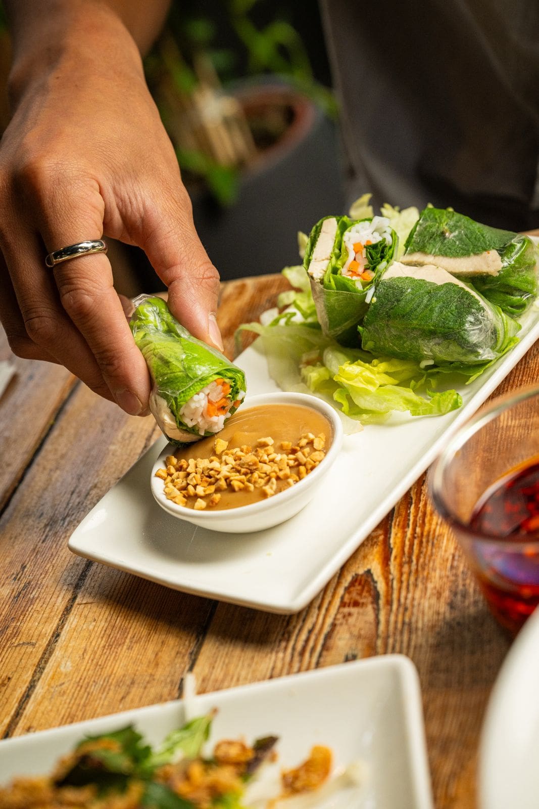 A hand dips a fresh spring roll filled with vegetables into a small bowl of peanut sauce, with more rolls and lettuce on a white plate nearby, set on a wooden table.