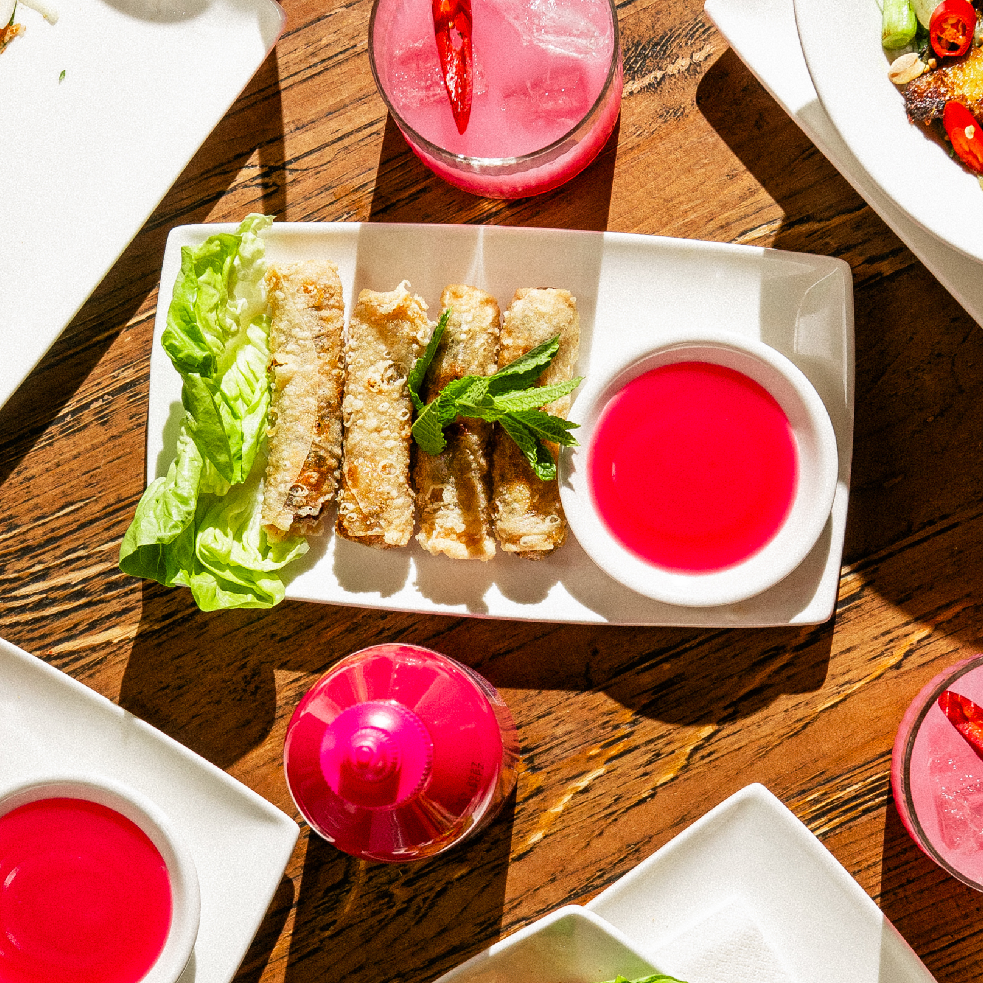 Rectangular plate with crispy spring rolls, lettuce, mint, and bright pink dipping sauce. Pink drink and other plates are partially visible on a wooden table. Sunlight creates strong shadows.