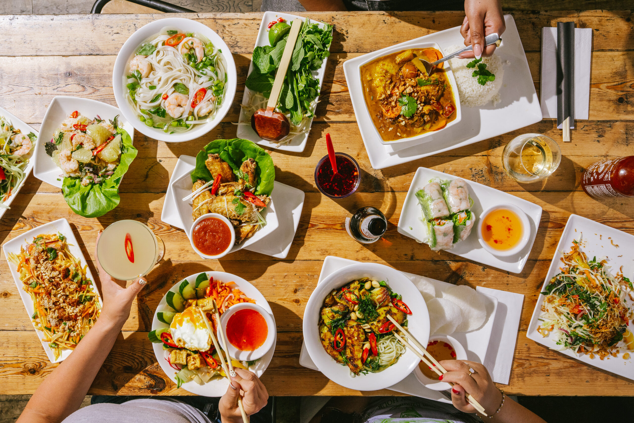 A table filled with various Vietnamese dishes, including noodles, spring rolls, salads, curry, and drinks, with people’s hands reaching for food and chopsticks in use.
