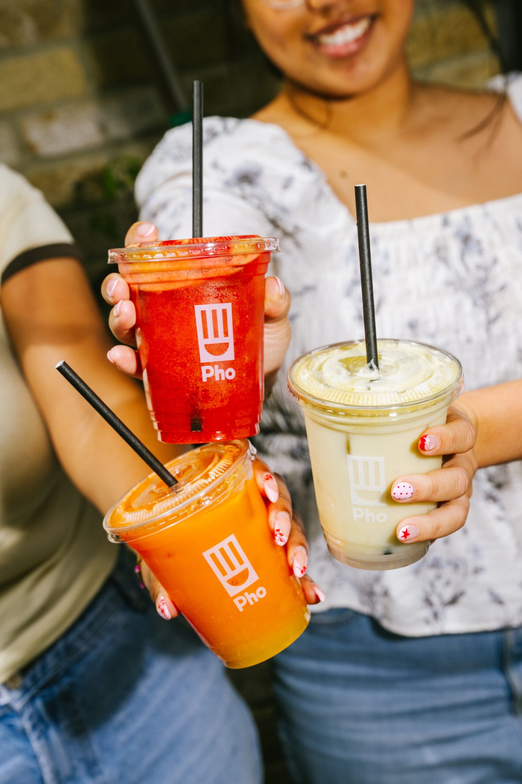Two people holding three colorful drinks with black straws in plastic cups labeled “Pho.” The drinks appear red, orange, and green. One person is smiling, and both have decorative nail art.