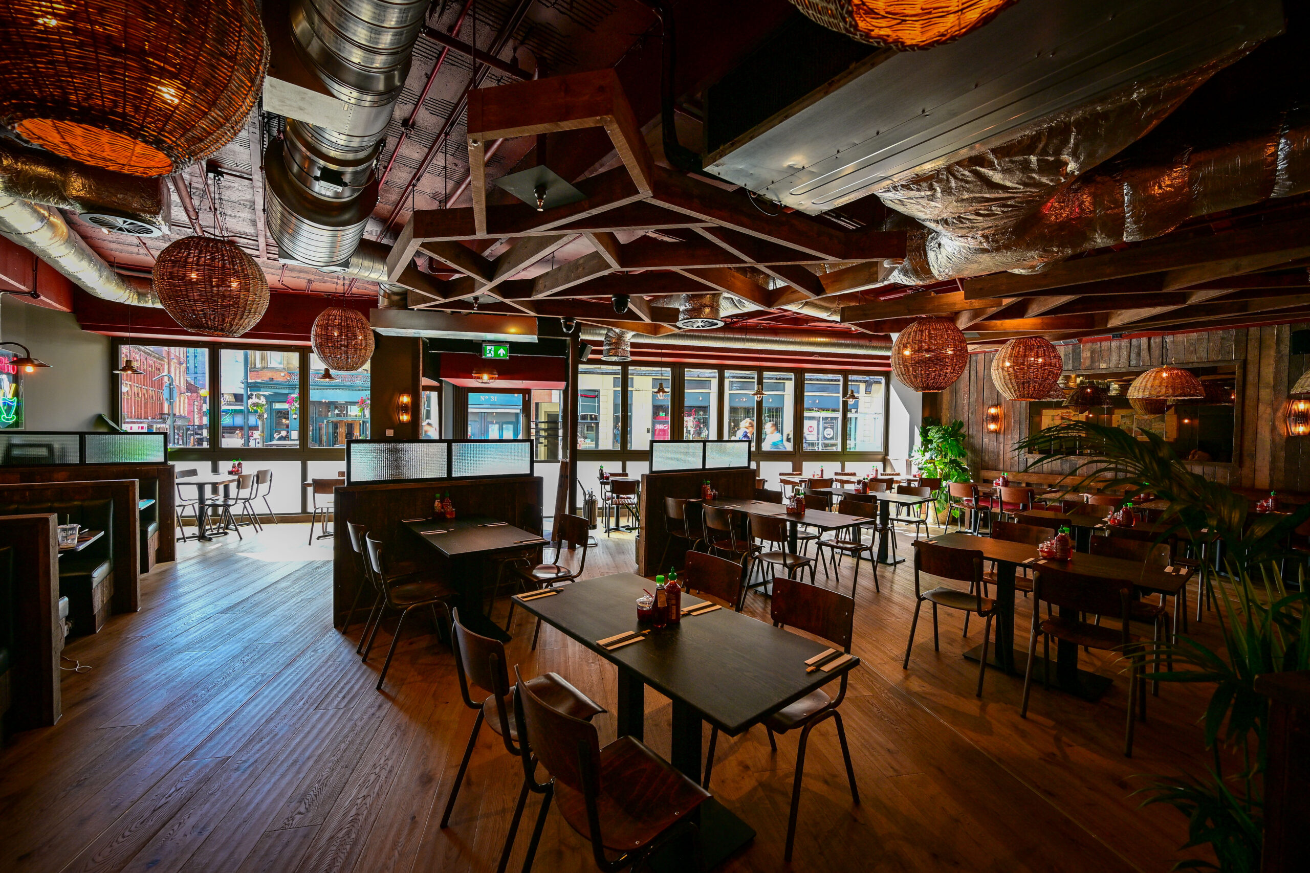 Spacious restaurant interior with wooden floors, tables, chairs, booth seating, hanging lights, exposed ceiling pipes, and sunlight streaming in through large windows.