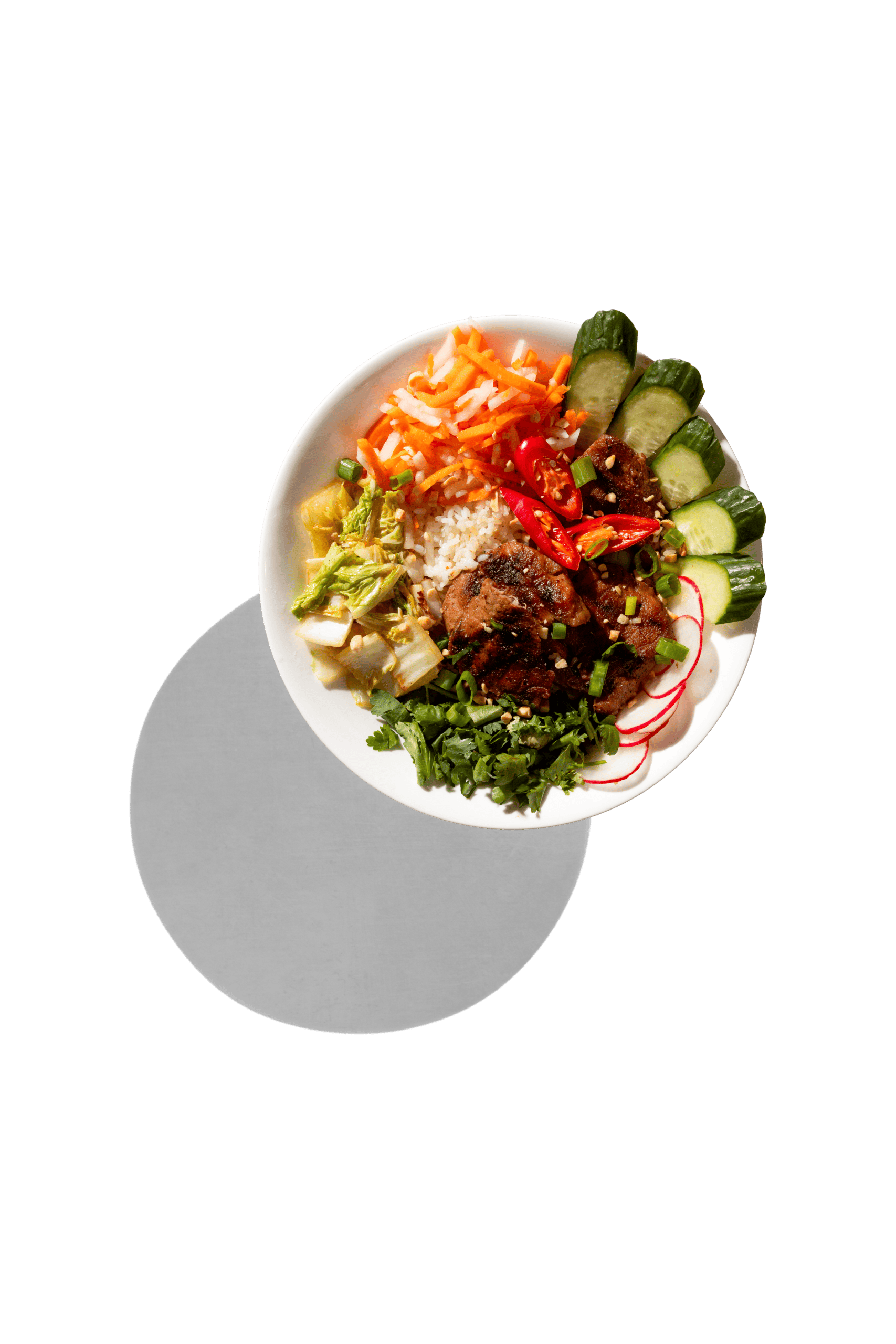 A bowl of rice topped with grilled meat, sliced cucumbers, radishes, carrots, greens, and chili peppers, photographed on a white background with a gray shadow.
